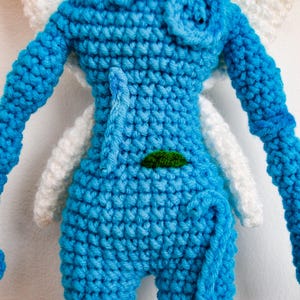 May include: A blue crocheted doll featuring a textured surface, a small green embroidered leaf detail on the torso, and large white crocheted wings extending from the back. The doll has long blue arms and a curled blue tail at the lower section.