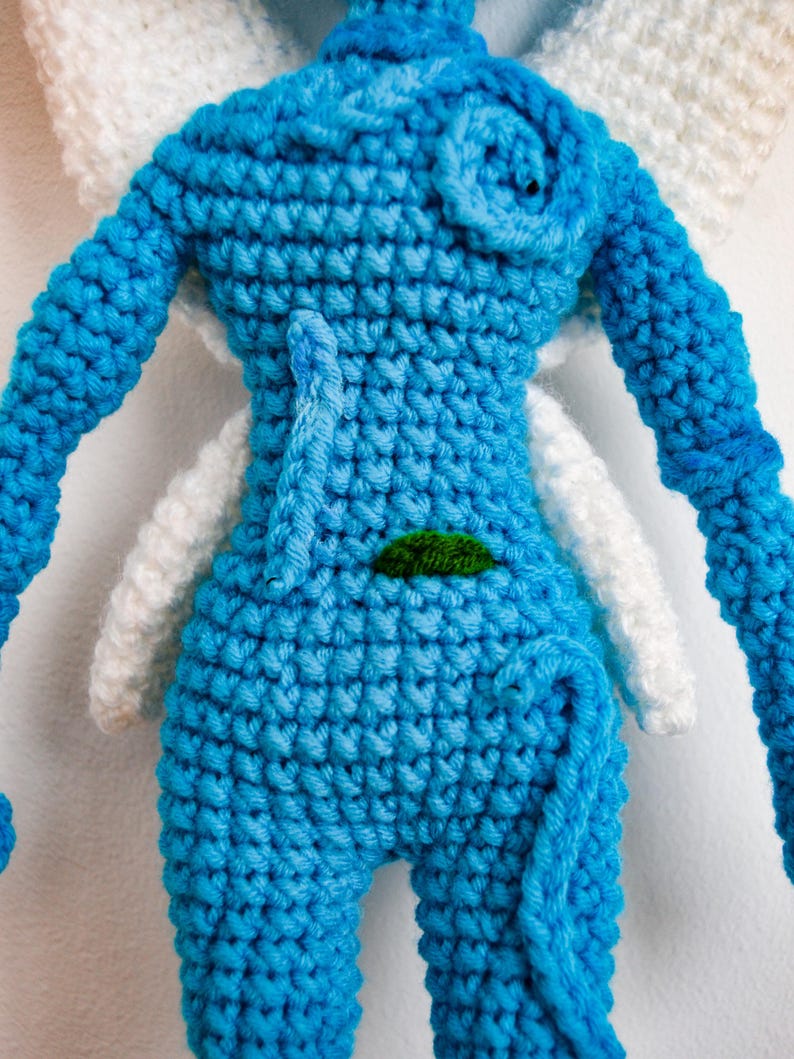 May include: A blue crocheted doll featuring a textured surface, a small green embroidered leaf detail on the torso, and large white crocheted wings extending from the back. The doll has long blue arms and a curled blue tail at the lower section.