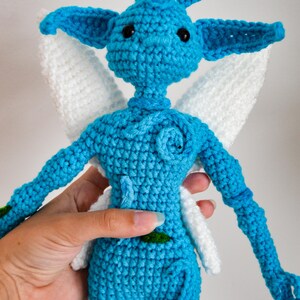 May include: A blue crocheted doll with large pointed ears, black bead eyes, and white wings. The figure features a textured surface with a small blue spiral detail on the chest and a tiny green leaf accent on the torso, crafted from yarn.