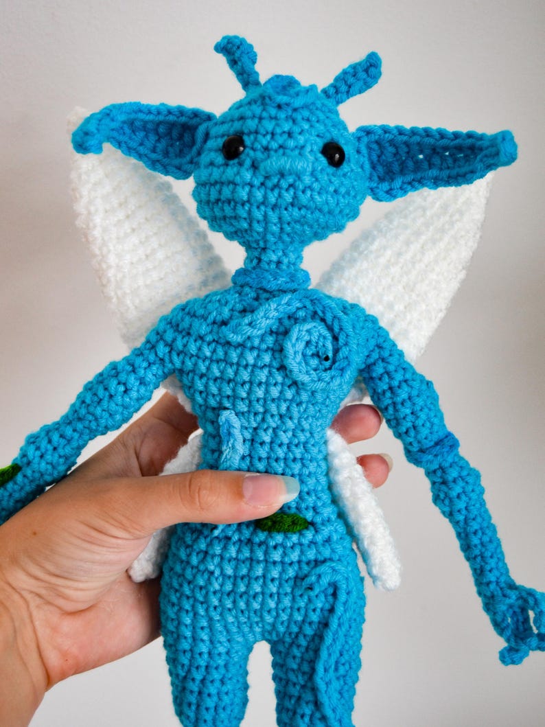 May include: A blue crocheted doll with large pointed ears, black bead eyes, and white wings. The figure features a textured surface with a small blue spiral detail on the chest and a tiny green leaf accent on the torso, crafted from yarn.