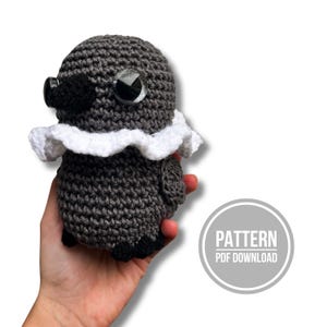 May include: A dark gray crocheted bird plush toy featuring a white ruffled collar, a black beak, and large black plastic safety eyes. The toy has a rounded body and small wings. A circular graphic in the corner displays the text PATTERN PDF DOWNLOAD.