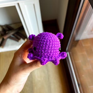 Op de afbeelding: Een kleine, levendig paarse octopus gemaakt van garen met een haaktechniek. Het knuffeltje heeft een ronde kop, een getextureerd oppervlak en kleine, gekrulde tentakels die vanaf de basis uitsteken, met twee zwarte, glanzende kraaloogjes.