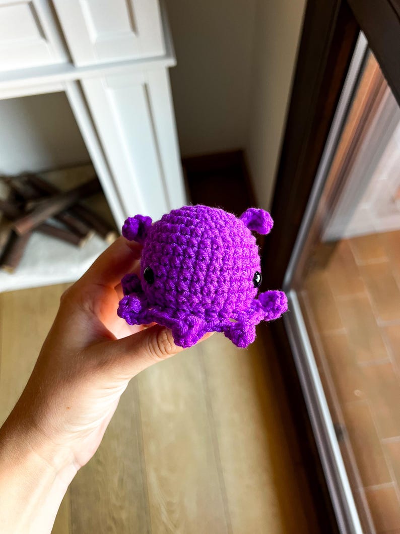 Op de afbeelding: Een kleine, levendig paarse octopus gemaakt van garen met een haaktechniek. Het knuffeltje heeft een ronde kop, een getextureerd oppervlak en kleine, gekrulde tentakels die vanaf de basis uitsteken, met twee zwarte, glanzende kraaloogjes.
