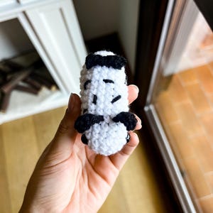 Op de afbeelding: Een klein, wit, gehaakt knuffeltje in de vorm van een zeeslak, met zwarte garenaccenten aan de boven- en zijkanten en twee kleine, ronde, zwarte plastic oogjes aan de voorzijde van het handgemaakte, getextureerde lichaam van textiel.