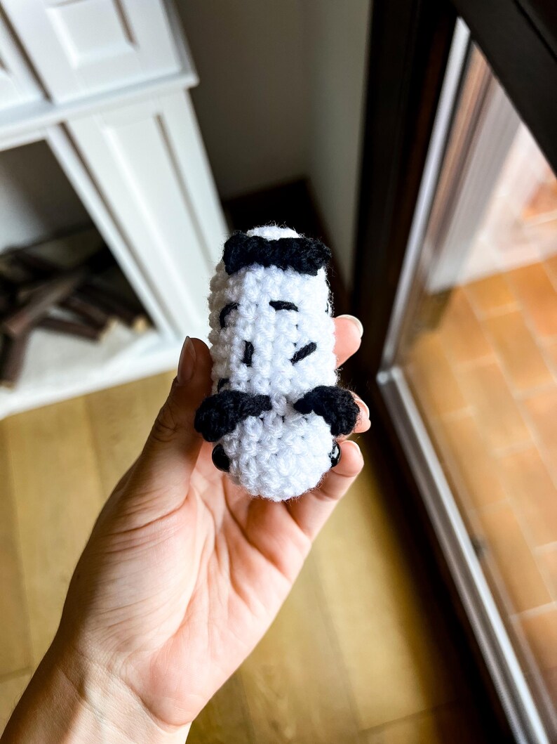 Op de afbeelding: Een klein, wit, gehaakt knuffeltje in de vorm van een zeeslak, met zwarte garenaccenten aan de boven- en zijkanten en twee kleine, ronde, zwarte plastic oogjes aan de voorzijde van het handgemaakte, getextureerde lichaam van textiel.