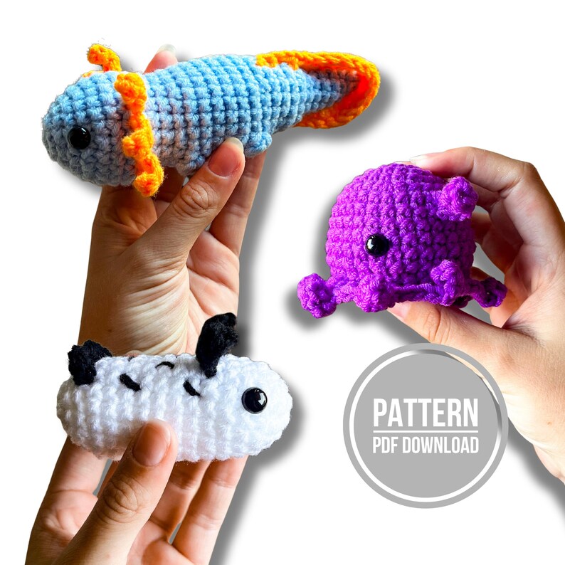 Op de afbeelding: Drie gehaakte knuffels waaronder een lichtblauwe axolotl met oranje kieuwen, een paarse octopus met kleine tentakels en een witte zeeslak met zwarte stippen. Een grijze badge met de tekst PATTERN PDF DOWNLOAD is zichtbaar.