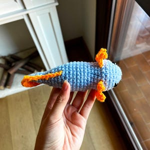 Op de afbeelding: Een lichtblauw gehaakte axolotl knuffel met feloranje uitwendige kieuwen en een bijpassende oranje rugvin. Het wezen heeft kleine zwarte kraaloogjes en een gestructureerde lichaamsvorm van garen met overal zichtbare steken.