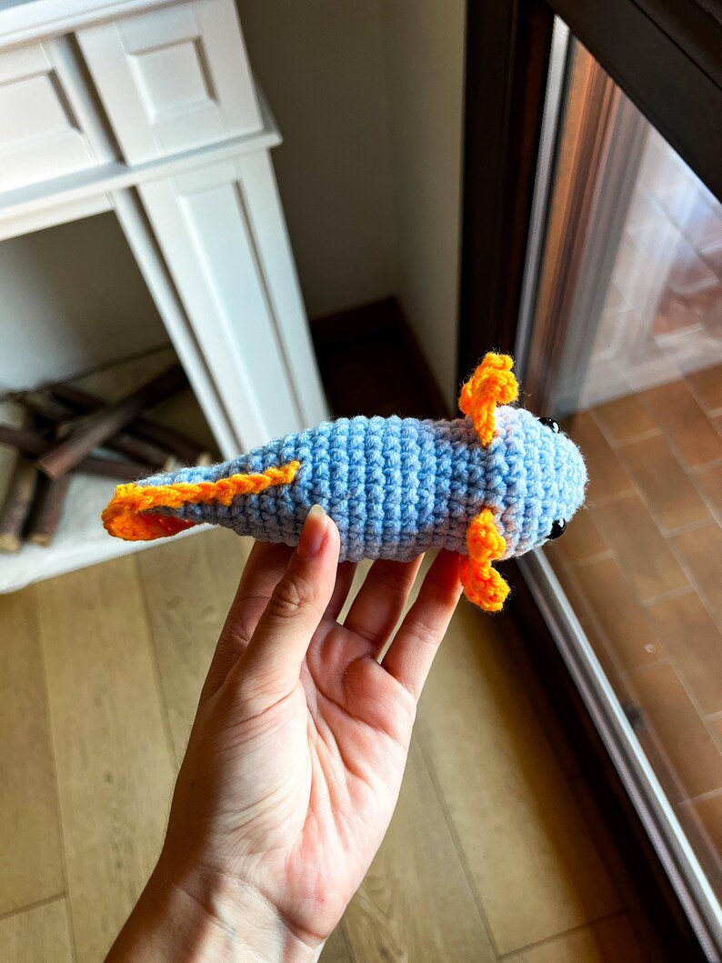 Op de afbeelding: Een lichtblauw gehaakte axolotl knuffel met feloranje uitwendige kieuwen en een bijpassende oranje rugvin. Het wezen heeft kleine zwarte kraaloogjes en een gestructureerde lichaamsvorm van garen met overal zichtbare steken.