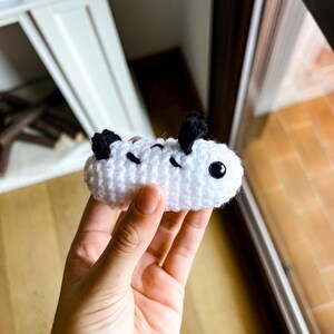 Op de afbeelding: Een klein, wit, gehaakt zeeslak-knuffeltje met een afgerond, cilindervormig lichaam. Het heeft twee zwarte, getextureerde, rechtopstaande rhinoforen aan de bovenkant, drie zwarte, gestikte diagonale markeringen op de zijkant en &eacute;&eacute;n zwart, glanzend oog.