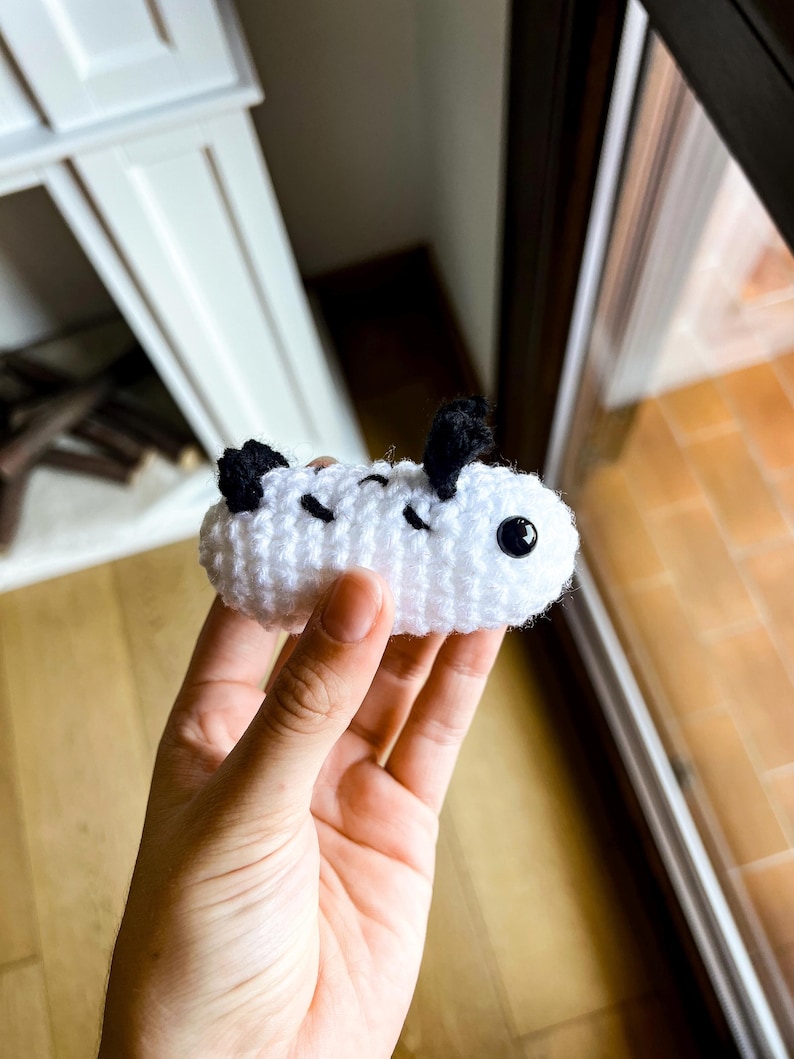 Op de afbeelding: Een klein, wit, gehaakt zeeslak-knuffeltje met een afgerond, cilindervormig lichaam. Het heeft twee zwarte, getextureerde, rechtopstaande rhinoforen aan de bovenkant, drie zwarte, gestikte diagonale markeringen op de zijkant en &eacute;&eacute;n zwart, glanzend oog.