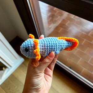 Op de afbeelding: Een klein, handgemaakt gehaakt axolotl-speeltje met een lichtblauw lichaam, feloranje uitwendige kieuwen en een bijpassende oranje staartvin. Het wezen heeft twee kleine, ronde, zwarte plastic ogen op het textuurrijke garen.