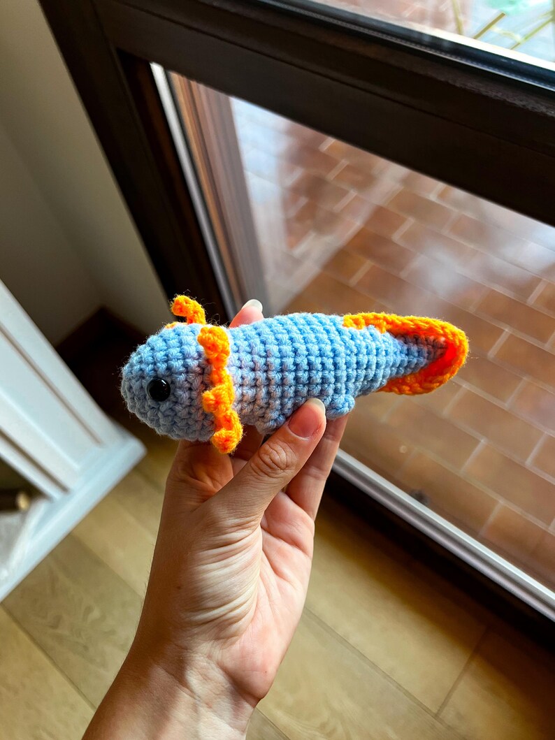 Op de afbeelding: Een klein, handgemaakt gehaakt axolotl-speeltje met een lichtblauw lichaam, feloranje uitwendige kieuwen en een bijpassende oranje staartvin. Het wezen heeft twee kleine, ronde, zwarte plastic ogen op het textuurrijke garen.