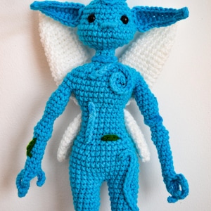 May include: A bright blue crocheted doll with large pointed ears, black bead eyes, and white crocheted wings. The figure features a small spiral detail on the chest, a thin tail, and small green leaf-like accents on the arm and torso.