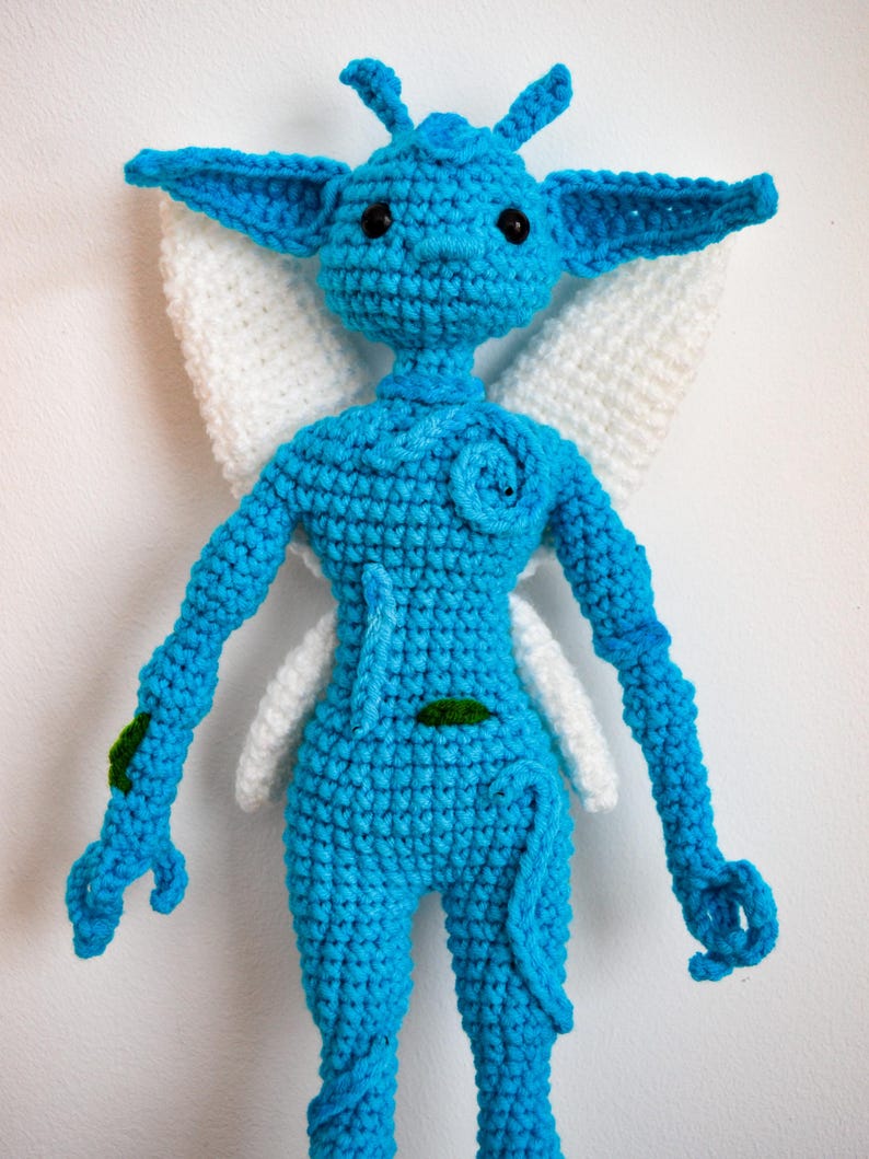 May include: A bright blue crocheted doll with large pointed ears, black bead eyes, and white crocheted wings. The figure features a small spiral detail on the chest, a thin tail, and small green leaf-like accents on the arm and torso.