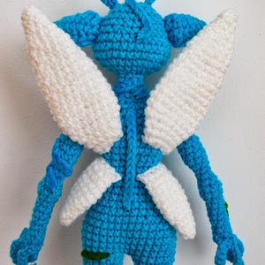 May include: A blue crocheted doll featuring a rounded head with two antennae, long arms, and two pairs of white crocheted wings attached to the back. The figure has a textured yarn surface and includes small green accents on the legs and arms.