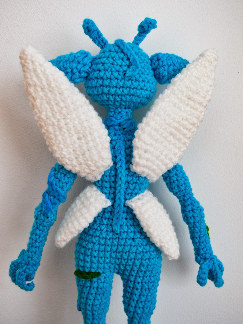 May include: A blue crocheted doll featuring a rounded head with two antennae, long arms, and two pairs of white crocheted wings attached to the back. The figure has a textured yarn surface and includes small green accents on the legs and arms.