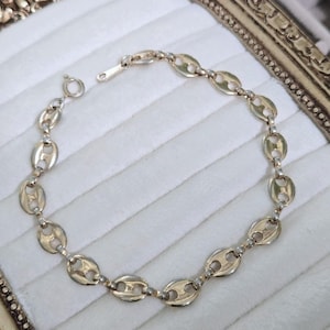May include: A gold-toned bracelet featuring a series of interconnected, oval-shaped links. The links have a distinctive design, resembling coffee beans. The bracelet is displayed on a white, textured surface, with a clasp visible at one end.