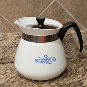 May include: A white teapot with a black lid and handle, featuring a metallic silver band around the upper section. The body of the teapot is decorated with a blue floral pattern consisting of small flowers and leaves centered on the front side.