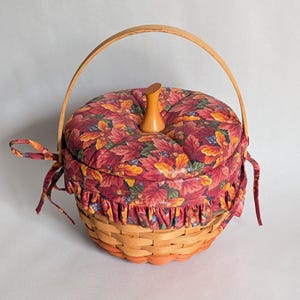 May include: A round woven wooden basket featuring a fabric cover with a red, orange, and yellow autumn leaf pattern. The lid has a central wooden knob, a ruffled fabric edge, and side ties. A curved wooden handle arches over the top of the basket.