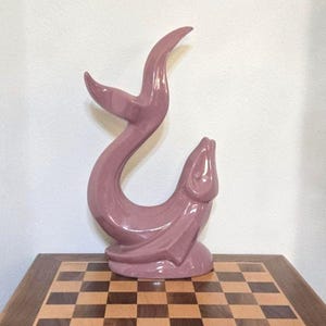 Puede incluir: Una escultura de cerámica malva de una criatura marina estilizada, posiblemente un delfín o una ballena, con un cuerpo y una cola curvados. La escultura se exhibe sobre una mesa de ajedrez de madera. La escultura mide aproximadamente 30 cm de altura.