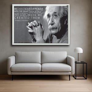 Może przedstawiać: Oprawiony czarno-biały portret Alberta Einsteina z cytatem: "WE CANNOT SOLVE OUR PROBLEMS WITH THE SAME THINKING WE USED WHEN WE CREATED THEM." Grafika wisi nad szarą sofą, z małym stolikiem i lampą z boku.