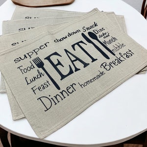 May include: Set of four beige linen placemats with black text and a fork and spoon graphic. The text reads "supper chow down snack food lunch feast EAT dine take-out munch nibble dinner homemade breakfast".