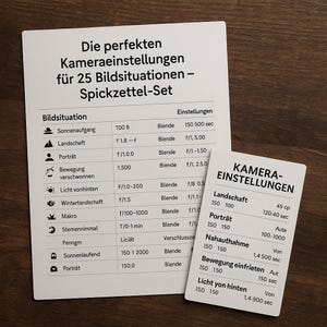 May include: Two white cards with German text on a wooden surface. The larger card lists camera settings for 25 situations, including landscape and portrait. The smaller card has camera settings for landscape, portrait, close-up, and backlighting.