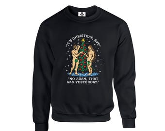 May include: A black crewneck sweatshirt featuring a pixel art graphic of Adam and Eve standing by a decorated Christmas tree. The text reads "IT'S CHRISTMAS, EVE" above the tree and "NO ADAM, THAT WAS YESTERDAY." below the figures.