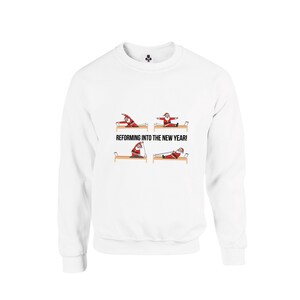 May include: A white crewneck sweatshirt featuring a graphic print of Santa Claus performing various Pilates exercises on a reformer machine. The text REFORMING INTO THE NEW YEAR! is printed in black capital letters below the illustrations.