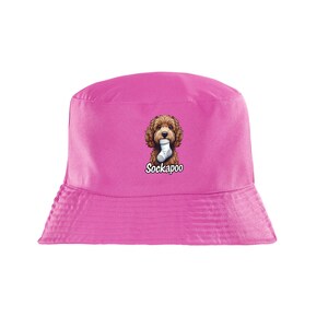 May include: A bright pink bucket hat featuring a centred graphic of a brown curly-haired dog holding a white sock in its mouth. Below the dog illustration, the text Sockapoo is printed in a bold, white, sans-serif font with a black outline.