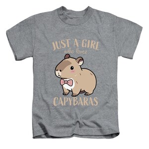 May include: A gray t-shirt featuring a cartoon capybara wearing a pink bow tie. The text "JUST A GIRL who loves CAPYBARAS" is printed above and below the capybara illustration. The shirt is designed for kids.