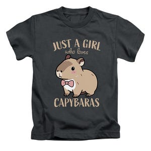 May include: Dark gray t-shirt featuring a cartoon capybara with a pink bow tie. The text reads "JUST A GIRL who loves CAPYBARAS" in a light brown color. The shirt is a crew neck style.