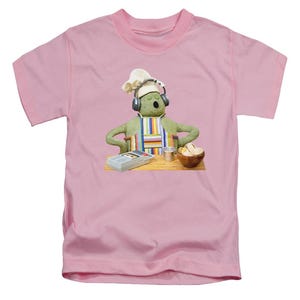 May include: A pale pink t-shirt with a cartoon chef character. The chef has green skin, a white hat, and headphones. The chef wears a striped apron and stands behind a wooden table with cooking utensils and ingredients.