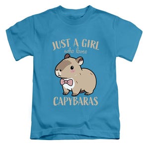 May include: A light blue t-shirt featuring a cartoon capybara with a pink bow tie. The text reads "JUST A GIRL who loves CAPYBARAS" in a light brown color. The shirt is designed for children.