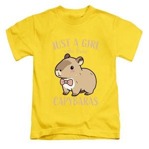 May include: A yellow t-shirt featuring a cartoon capybara wearing a pink bow tie. The text "JUST A GIRL who loves CAPYBARAS" is printed above and below the capybara illustration. The shirt is made of a soft material.