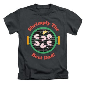 May include: Dark grey t-shirt with a graphic of a barbecue grill with shrimp and the text "Shrimply The Best Dad!". The design features pink shrimp, a green grill, and orange and yellow accents.