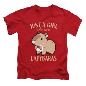 May include: Red t-shirt featuring a cartoon capybara wearing a pink bow tie. The text "JUST A GIRL who loves CAPYBARAS" is printed above and below the capybara illustration. The shirt is made of a soft material.