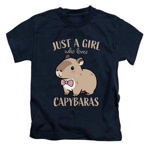 May include: Navy blue t-shirt featuring a cartoon capybara wearing a pink bow tie. The text "JUST A GIRL who loves CAPYBARAS" is printed above and below the capybara illustration. The shirt is designed for children.