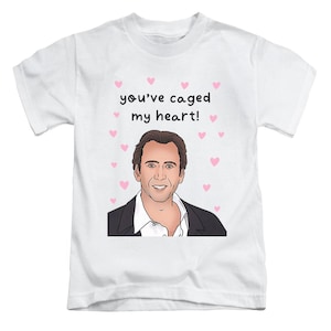 May include: White t-shirt with a cartoon illustration of a smiling man, the text "you've caged my heart!" and pink hearts. The man is wearing a black suit jacket and a white shirt.