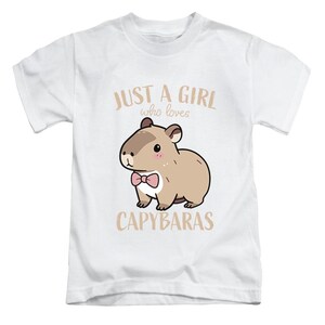 May include: White t-shirt featuring a cartoon capybara wearing a pink bow tie. The text reads "JUST A GIRL who loves CAPYBARAS" in a light brown color. The design is centered on the front of the shirt.