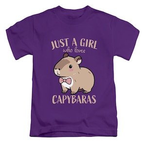 May include: Purple t-shirt featuring a cartoon capybara with a pink bow tie. The text reads "JUST A GIRL who loves CAPYBARAS" in a light brown color. The design is centered on the front of the shirt.