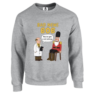 May include: A heather grey crewneck sweatshirt featuring a cartoon graphic of a doctor speaking to a nutcracker. The text reads BAD NEWS BOB and You've got a nut allergy. The doctor wears a white coat, and the nutcracker wears a red uniform.