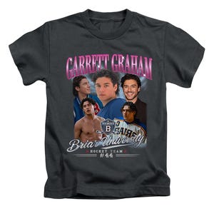 May include: A charcoal grey short-sleeve t-shirt featuring a graphic print of Garrett Graham. The design includes multiple photos of a person, the text "GARRETT GRAHAM" in pink, "Briar University" in white, and "HOCKEY TEAM #44" at the bottom.