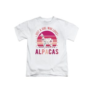 May include: A white short-sleeve t-shirt featuring a graphic of an alpaca filled with floral patterns. Behind the alpaca is a pink and orange sunset circle. The text reads JUST A GIRL WHO LOVES ALPACAS in pink and orange lettering.