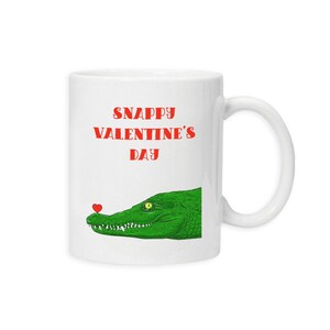May include: White ceramic mug featuring a green alligator graphic with a red heart on its nose. The mug has the text "Snappy Valentine's Day" in red. The mug has a white handle.