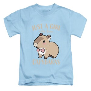 May include: Light blue t-shirt featuring a cartoon capybara wearing a pink bow tie. The text "JUST A GIRL who loves CAPYBARAS" is printed above and below the capybara illustration. The shirt is designed for children.