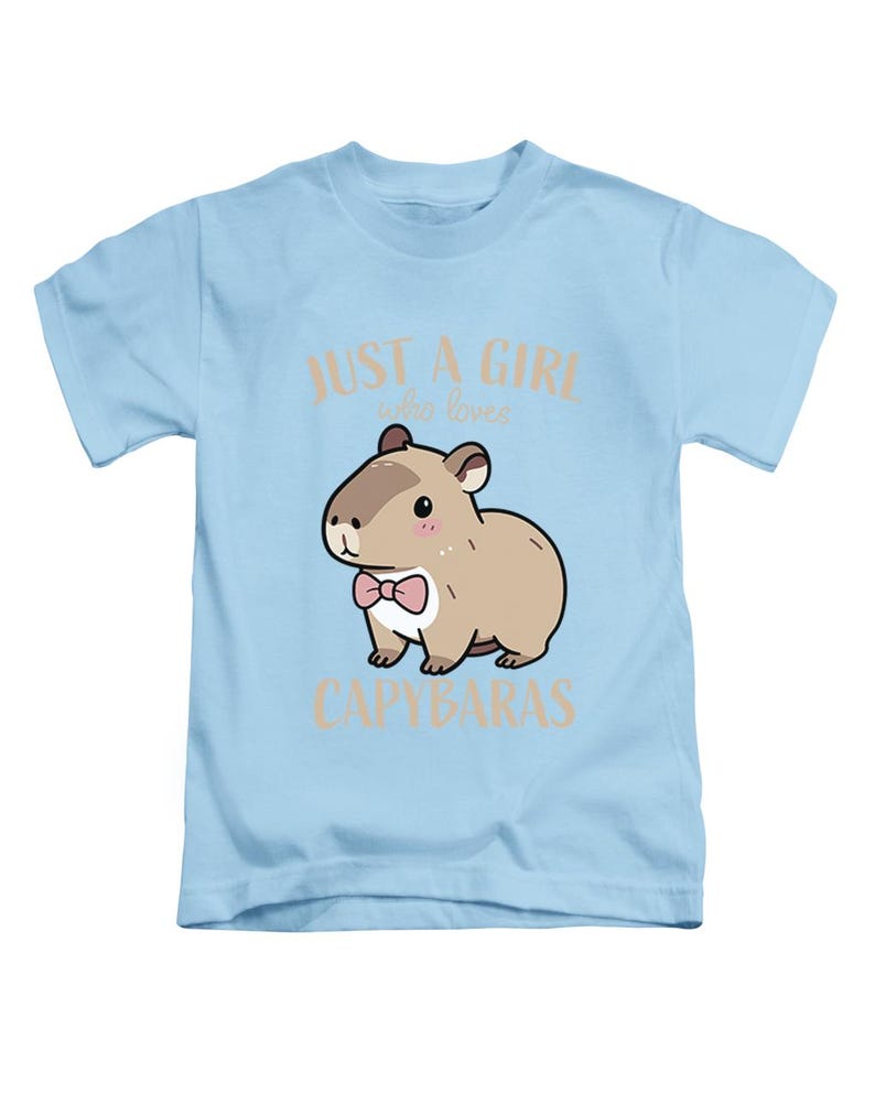 May include: Light blue t-shirt featuring a cartoon capybara wearing a pink bow tie. The text "JUST A GIRL who loves CAPYBARAS" is printed above and below the capybara illustration. The shirt is designed for children.
