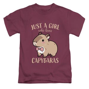 May include: A maroon t-shirt featuring a cartoon capybara wearing a pink bow tie. The text "JUST A GIRL who loves CAPYBARAS" is printed above and below the capybara illustration. The shirt is made of a soft material.