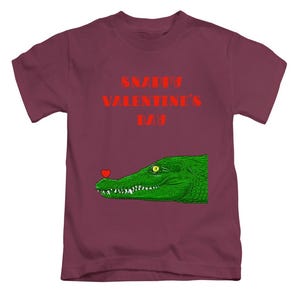 May include: A maroon t-shirt with a green alligator graphic and a red heart on its nose. The text "SNAPPY VALENTINE'S DAY" is printed in red above the alligator. This casual t-shirt is suitable for everyday wear.