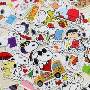 Peut inclure: Une collection d'autocollants colorés mettant en vedette Snoopy et d'autres personnages de Peanuts. Les autocollants représentent Snoopy dans diverses poses, notamment avec des lunettes d'aviateur, tenant un cœur et conduisant un bus scolaire jaune. Lucy et Woodstock sont également présents.